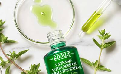 Flasche mit Kiehl's Cannabis Sativa Seed Oil, umgeben von Krautern und Tropfer, auf marmoriertem Hintergrund.