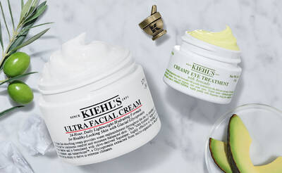 Zwei Kiehls Hautpflegeprodukte: Ultra Facial Cream und Creamy Eye Treatment, umgeben von Olivenzweigen und Avocado.