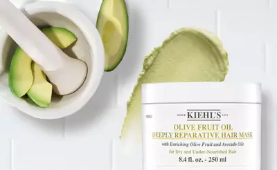 Kiehls Haarmaske mit Oliven- und Avocadool, umgeben von einer halben Avocado und einer Morser.