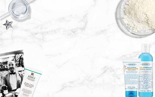 Kiehl's Produkte (Reiniger, Adstringens) und historische Elemente (Mann, Messbecher) auf weißem Marmorhintergrund.