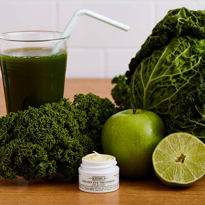 Gruner Smoothie mit Strohhalm, frischem Grunkohl, Apfel, Limette und einer Kiehls Augencreme auf einem Holztisch.