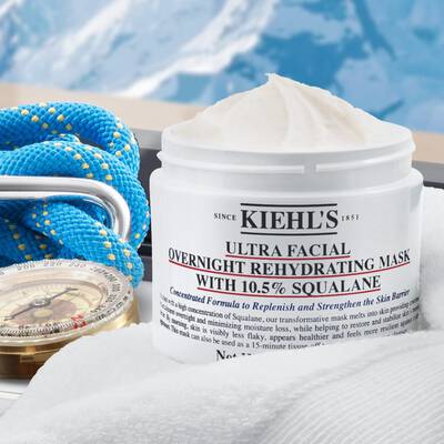 Ein Tiegel Kiehl's Ultra Facial Overnight Rehydrating Mask mit einer cremigen Textur, umgeben von blauen Seilen und einer Kompassuhr auf einem weißen Handtuch, mit Bergen im Hintergrund.