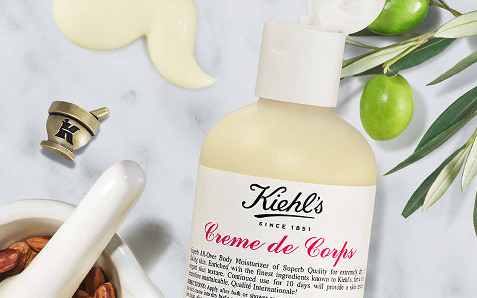 Alt-Text: "Produktauswahl von Kiehl's, einschließlich Creme de Corps und Hautpflegeole, umgeben von naturlichen Zutaten wie Vanille und Oliven."