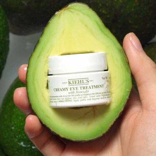 Ein Kiehl's Creamy Eye Treatment with Avocado ist in einer aufgeschnittenen Avocado platziert und wird von einer Hand gehalten.