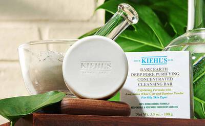 Ein Kiehls Reinigungsbar aus weißem Ton und Bambuspulver, umgeben von grunen Pflanzen und einer Verpackung im Hintergrund.