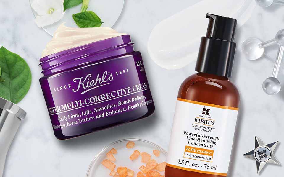 Ein stilvolles Arrangement von Hautpflegeprodukten auf einem Marmortisch, einschließlich einer lila Creme von Kiehl's und einem braunen Serum von Kiehl's, umgeben von grunen Blattern und einem Spiegel.