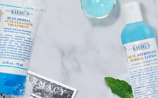 Alt-Text: Eine flache Anordnung von Kiehl's Hautpflegeprodukten auf einem marmorierten Hintergrund, einschließlich einer Tube mit Gesichtscreme, einer Flasche mit Lotion und frischen Krautern.