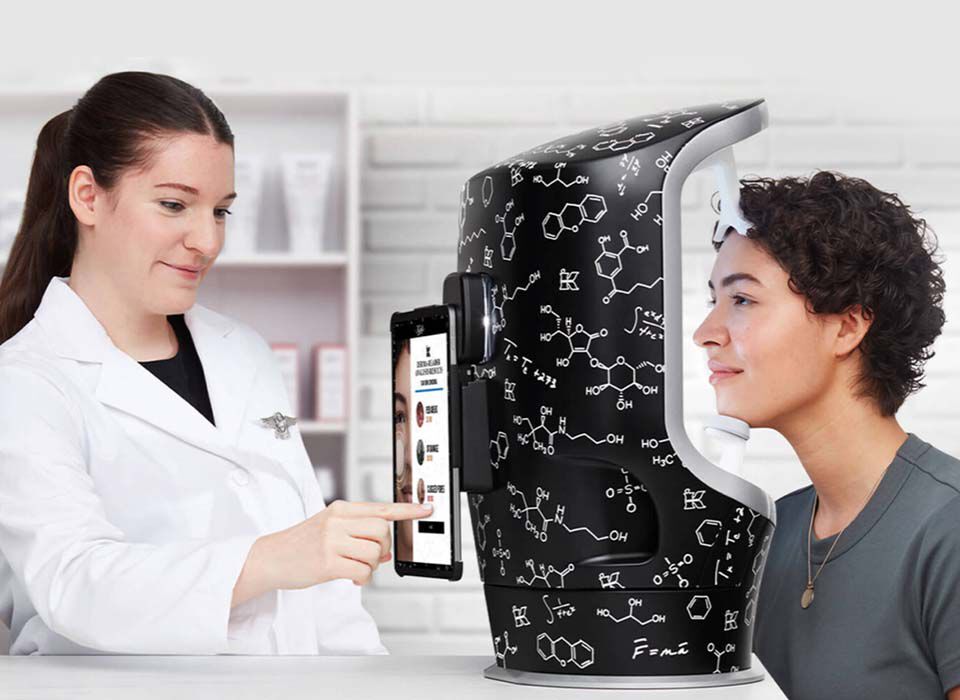 Eine Fachkraft erklart einer Person, die neben einem innovativen Gerat steht, wie man Hautanalysen durchfuhrt. Das Gerat hat eine moderne, wissenschaftlich anmutende Oberflache.