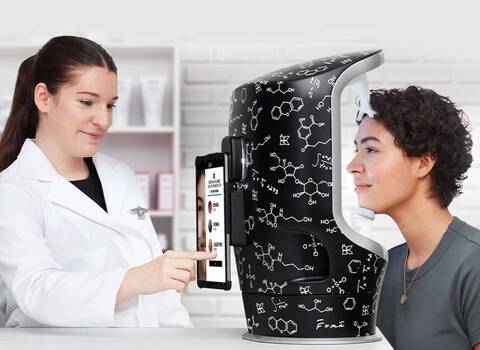 Eine Fachkraft erklart einer Person, die neben einem innovativen Gerat steht, wie man Hautanalysen durchfuhrt. Das Gerat hat eine moderne, wissenschaftlich anmutende Oberflache.