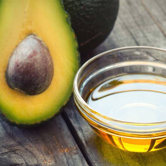 Halbe Avocado mit Kern, ganze Avocado und Schale mit Avocadool auf rustikalem Holztisch.