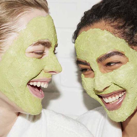 Zwei lachende Frauen tragen grune Gesichtsmasken und schauen sich an, genießen offensichtlich einen entspannten Beauty-Moment.
