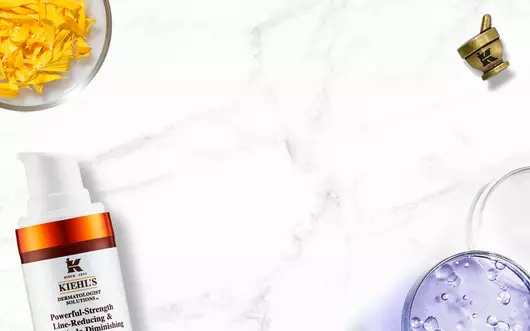 Alt-Text: Eine flache Anordnung von Kiehl's Hautpflegeprodukten auf einem marmorierten Hintergrund, einschließlich einer Tube mit Gesichtscreme, einer Flasche mit Lotion und frischen Krautern.