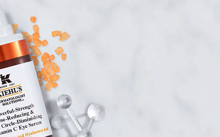 Alt-Text: Auf einem marmorierten Hintergrund sind zwei Hautpflegeprodukte von Kiehl's zu sehen. Links befindet sich ein Serum mit orangefarbenen Inhaltsstoffen, rechts ein Midnight Recovery Eye Balsam, umgeben von dekorativen Akzenten wie Lavendel und kleinen silbernen Objekten.