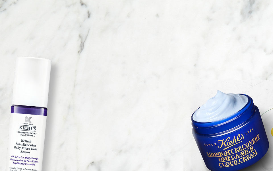 Alt-Text: Eine flache Anordnung von Kiehl's Hautpflegeprodukten auf einem marmorierten Hintergrund, einschließlich einer Tube mit Gesichtscreme, einer Flasche mit Lotion und frischen Krautern.