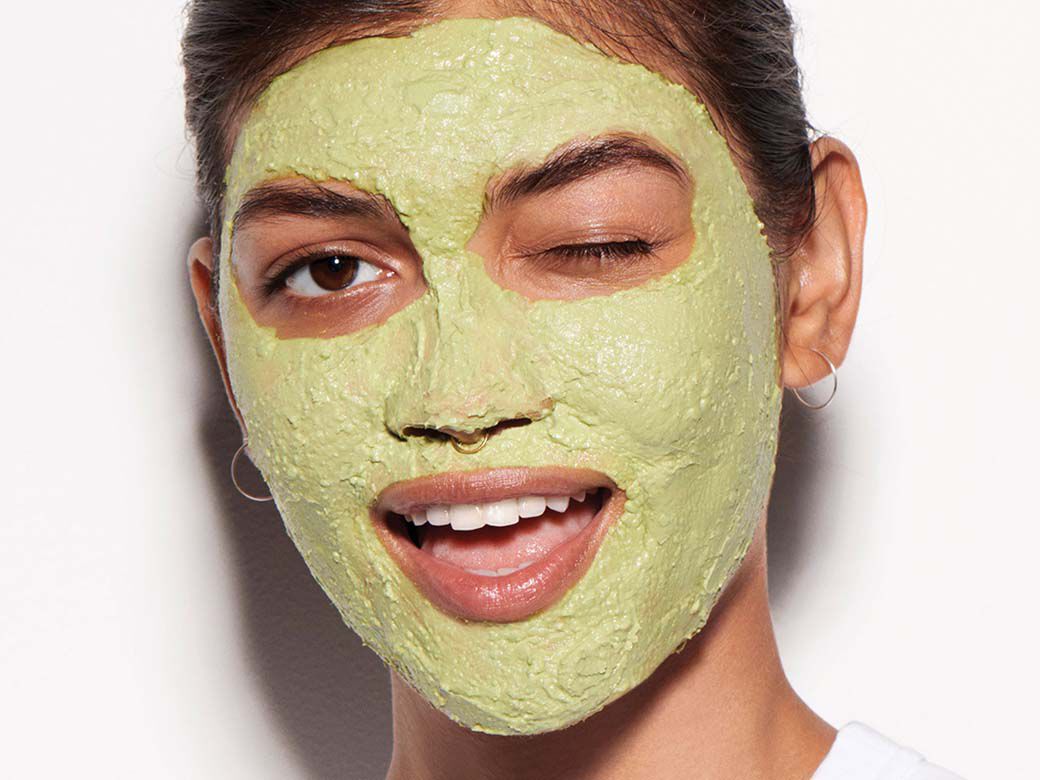 Eine Frau mit gruner Gesichtsmaske, einem Zwinkern und einem breiten Lacheln blickt in die Kamera.