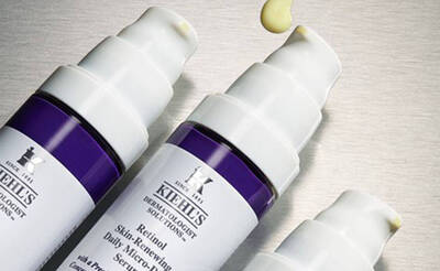 Drei Flaschchen Kiehl's Retinol-Serum mit einer Tropfen an der Spitze, vor einem metallischen Hintergrund.