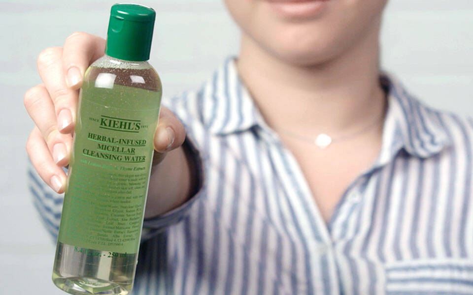 Eine Frau mit lockigen Haaren halt ein kosmetisches Produkt in der Hand, umgeben von Lavendel und einem Reinigungsgel von Kiehl's auf einem marmorierten Hintergrund.