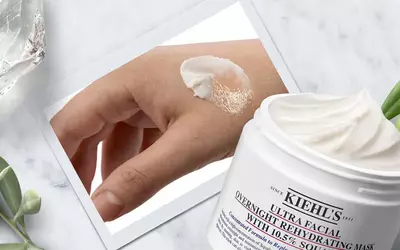 Handedemonstration einer Kiehl's Gesichtsmaske auf einer Hautpartie, umgeben von Pflanzen und Produktverpackung.