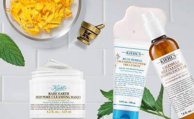 Alt text: A flat lay of Kiehl's skincare products including a jar of Rare Earth Deep Pore Cleansing Masque, a tube of Blue Herbal Blemish Cleanser Treatment, and a bottle of Calendula Deep Cleansing Face Wash, accompanied by yellow flower petals and a mortar and pestle.