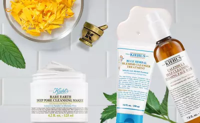 Alt text: A flat lay of Kiehl's skincare products including a jar of Rare Earth Deep Pore Cleansing Masque, a tube of Blue Herbal Blemish Cleanser Treatment, and a bottle of Calendula Deep Cleansing Face Wash, accompanied by yellow flower petals and a mortar and pestle.