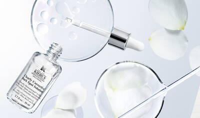 A flat lay of skincare products featuring a dropper bottle of Kiehl's Clearly Corrective Dark Spot Solution, surrounded by glass petri dishes with water droplets and white rose petals.