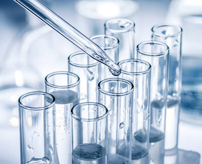 A pipette is dripping liquid into a series of clear test tubes filled with water, set against a blurred laboratory background.