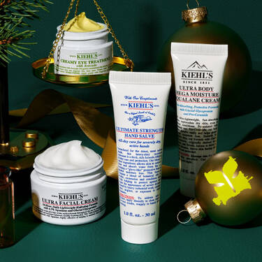 Eine festliche Anordnung von Kiehl’s Produkten, darunter Creamy Eye Treatment auf goldenen Waagschalen, Ultra Facial Cream, Ultimate Strength Hand Salve und Ultra Body Mega Moisture Squalane Cream, umgeben von grunen Ornamenten, Tannenzweigen und goldenen Bandern auf einem dunkelgrunen Hintergrund.