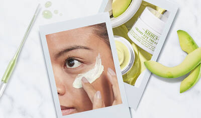 A close-up of a person applying a white cream under their eye with a smooth texture. In the background, there are two jars of skincare products and slices of green fruit, suggesting moisturizing ingredients.