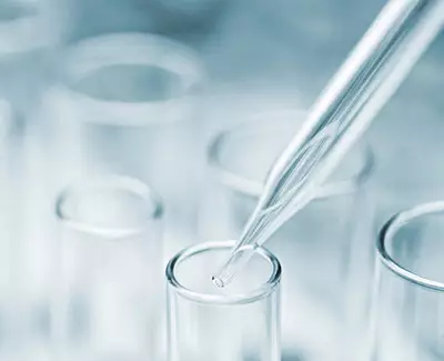 A close-up image of a glass pipette above a test tube, with a blurred laboratory background.