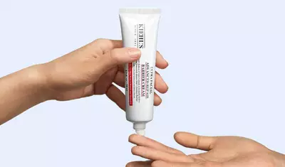A hand is holding a tube of Kiehl's Vitamin C cream, while another hand extends beneath it, ready to receive the product. The background is a soft, light blue.