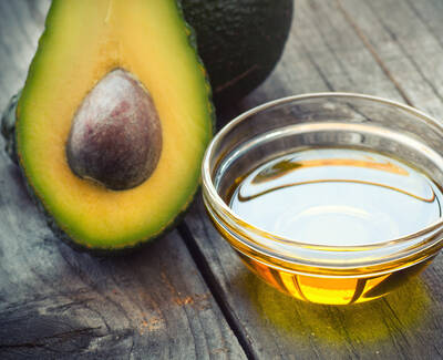 A halved avocado with a pit sits next to a small glass bowl filled with golden oil, all displayed on a wooden surface.