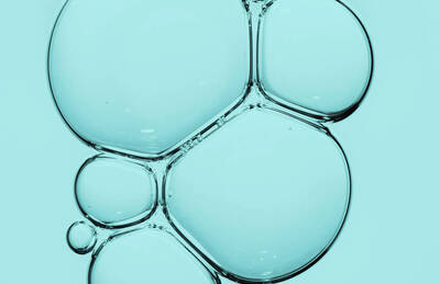 Close-up of translucent bubbles in a light blue background, varying in size and shape, creating a fluid and abstract appearance.