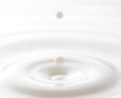A close-up image of a droplet falling into a smooth, white liquid, creating ripples as it impacts the surface.