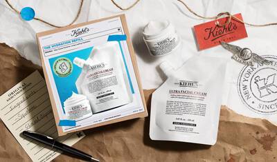 A flat lay of Kiehl's skincare products, featuring a box labeled "The Hydration Refill" alongside a pouch of Ultra Facial Cream, a small jar of the cream, a list of ingredients on a notepad, and a pen, all presented on crumpled brown paper.