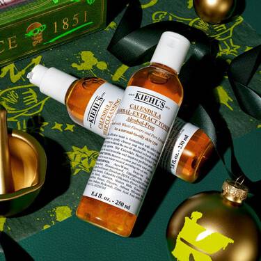 Kiehl’s Calendula Herbal-Extract Toner und Serum, arrangiert mit goldenen Weihnachtsornamenten und grunem Band auf einem festlichen, dunkelgrunen Hintergrund.