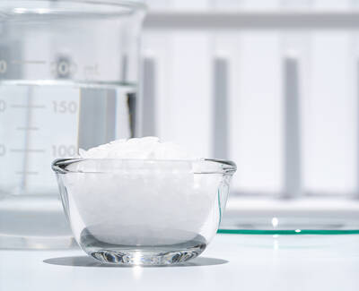 A clear glass bowl filled with white granules is placed next to a glass measuring beaker filled with water, all set against a light background.