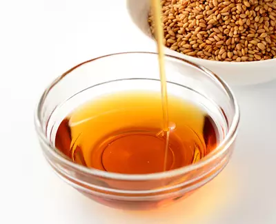 A glass bowl containing golden sesame oil, with oil being poured in, and a bowl of sesame seeds in the background.