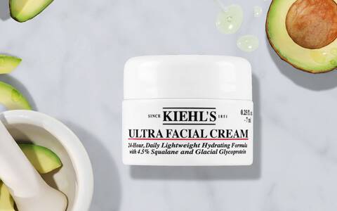 Produktbild der Kiehl's Ultra Facial Cream, leichte 24-Stunden-Feuchtigkeitsformel mit 4,5% Squalan und Gletscherprotein (7 ml), prasentiert auf grauem Marmorhintergrund mit frischen Avocadoscheiben und Serumtropfen.