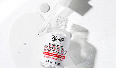 A clear bottle of Kiehl's Ultra Pure High-Potency Serum with 38% Glycolic Acid, resting on a white surface with some serum droplets beside it.