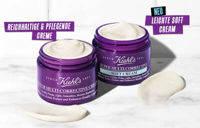 Two jars of Kiehl's Super Multi-Corrective Soft Cream, one open, showcasing a smooth, creamy texture. The jars are purple with white labels, and the text features German phrases as well as "NEU" (new). A small amount of cream is spread on the surface beside the jars.