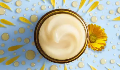 A swirl of creamy moisturizer in a brown jar, surrounded by yellow flower petals and droplets of water, on a light blue background.
