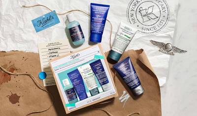 A flat lay of Kiehl's skincare products, including tubes and bottles, arranged around a gift set box and surrounded by a recycled paper backdrop, featuring the Kiehl's logo and a note detailing ingredients.