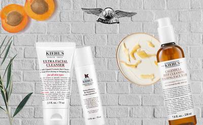A collection of Kiehl's skincare products displayed against a textured white brick background, including Ultra Facial Cleanser, a serum, and Calendula Deep Cleansing Foaming Face Wash, with apricots and botanical elements accenting the scene.