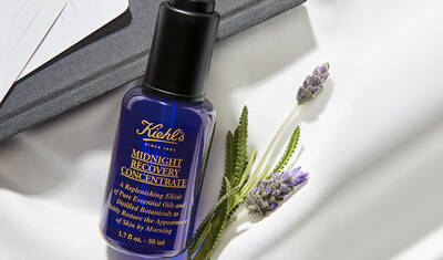 An elegant bottle of Kiehl's Midnight Recovery Concentrate alongside fresh lavender and a gray notebook, resting on a soft white surface.