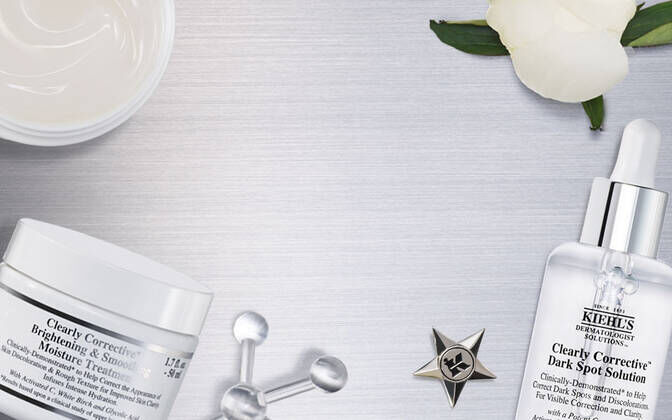 Produits de soins de la peau Kiehl's sur une surface en aluminium, comprenant un toner, un serum et une creme, entoures d'elements decoratifs comme des fleurs blanches.