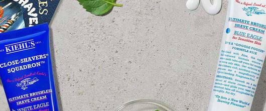 Une composition de cremes de rasage Kiehl's sur une surface grise, entourees de feuilles de menthe et d'aloe vera, avec un rasoir en metal.