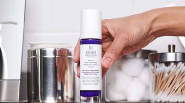 Un flacon de serum pour le visage de la marque Kiehl's, avec une main feminine le tenant. En arriere-plan, on voit des accessoires de salle de bain comme des cotons et des cure-dents.