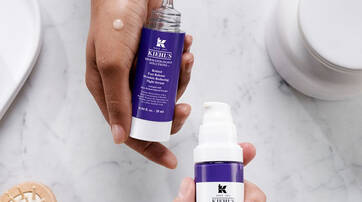 Deux flacons de serum de la marque Kiehl's, l'un avec un embout compte-gouttes et l'autre avec une pompe, poses sur une surface marbree. Une main tient l’un des flacons, tandis qu'une goutte de produit est visible sur l'autre.