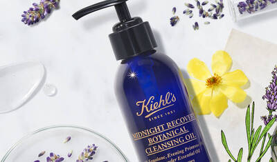 A bottle of Kiehl's Midnight Recovery Botanical Cleansing Oil is displayed with lavender flowers and petals. The bottle features a pump top and has a deep blue hue with gold lettering.