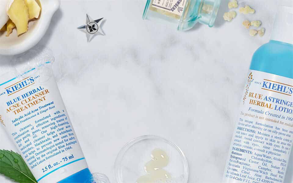 Produits de soin de la peau de la marque Kiehl's sur un fond marbre, incluant des flacons de lotion et des ingredients naturels comme des feuilles de menthe et des fleurs.